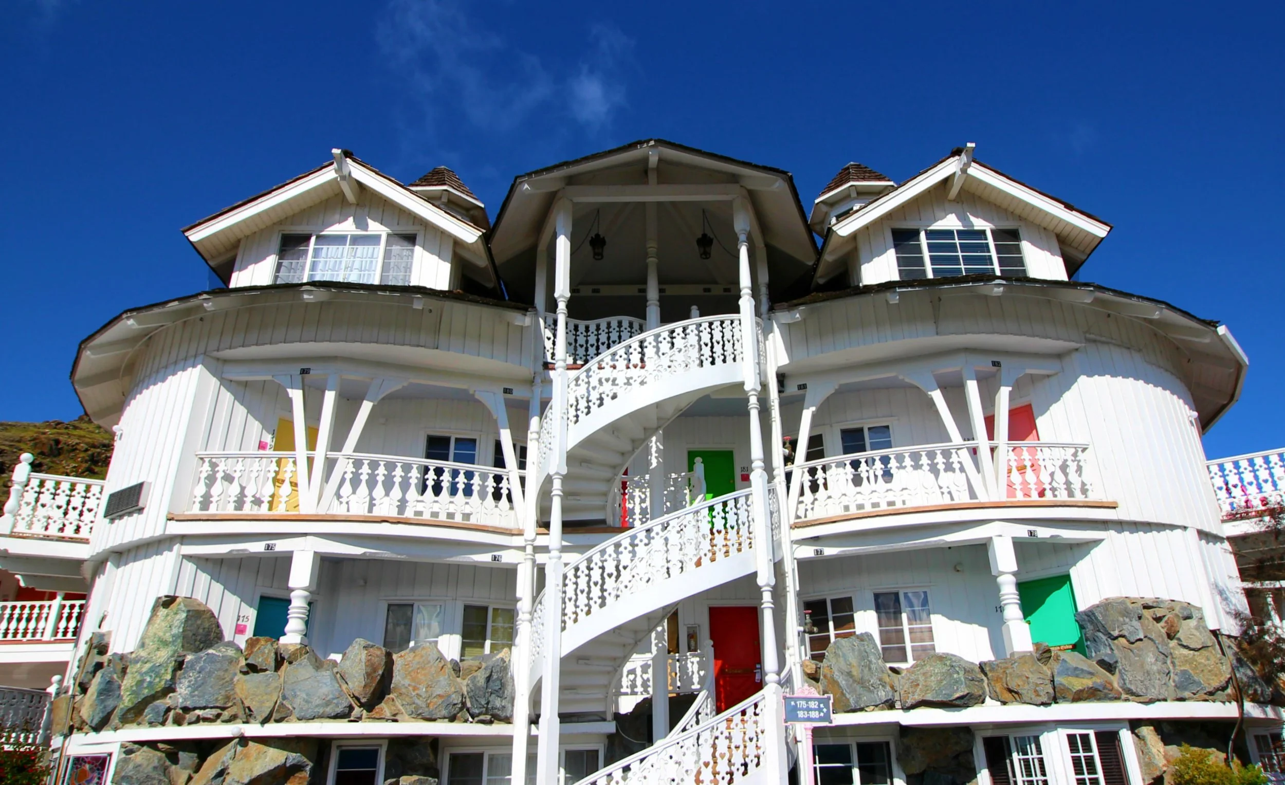 A Madonna Inn guest wing in daylight — white alpine balconies, a spiral staircase, and brightly colored room doors